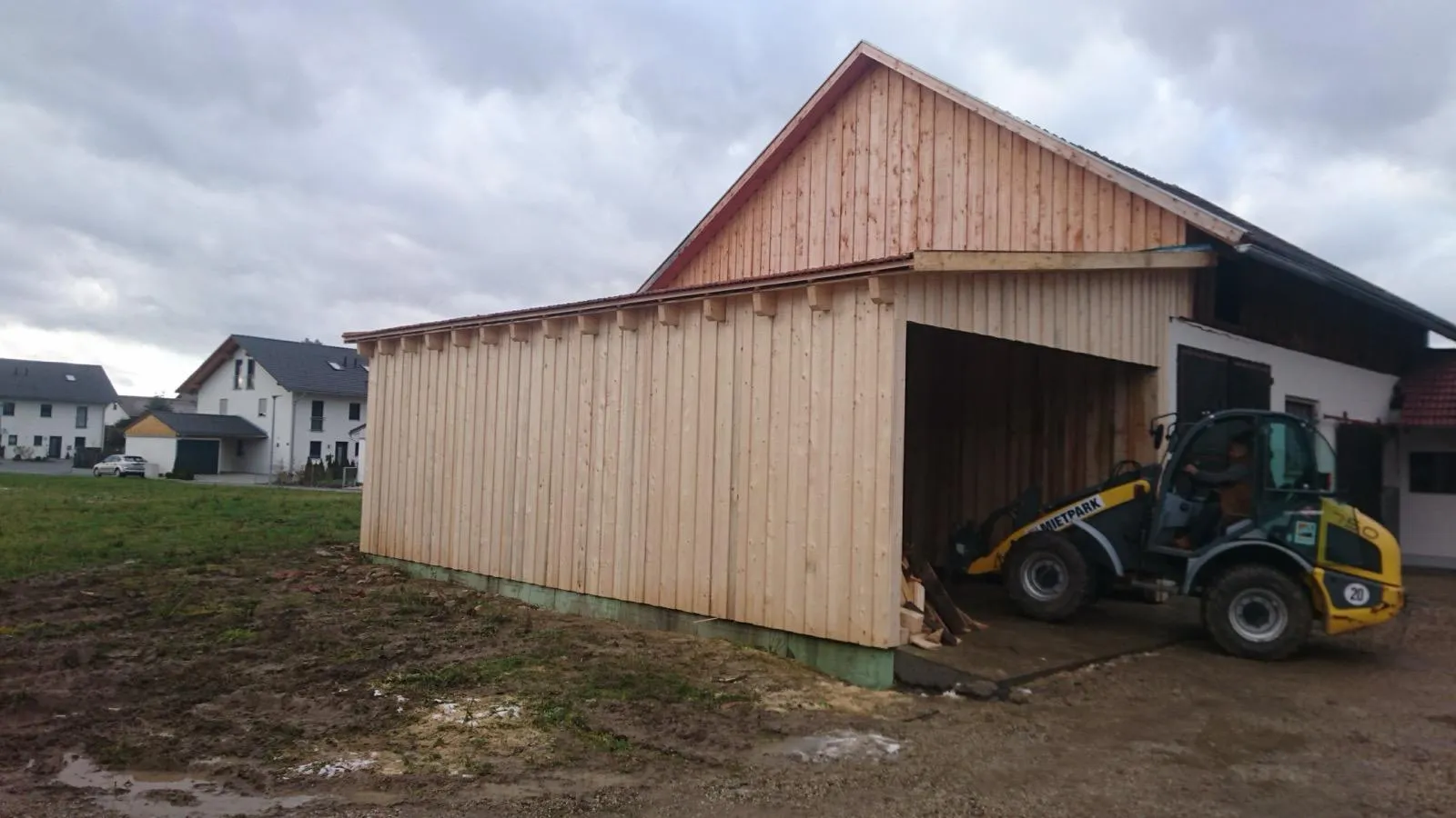 Drewniany garaż przy domu z otwartą wnęką wjazdową i konstrukcją dopasowaną do istniejącej elewacji.